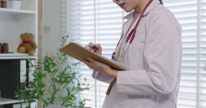 Closeup of asian female pediatrician in white coat writing notes on clipboard during medical checkup routine highlighting focus organization professionalism in pediatric healthcare practice