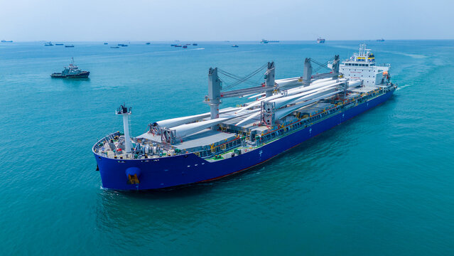 Large heavy lift cargo ship transporting massive wind turbine blades across the deep blue ocean. Global logistics for renewable energy and maritime shipping industry business concept.