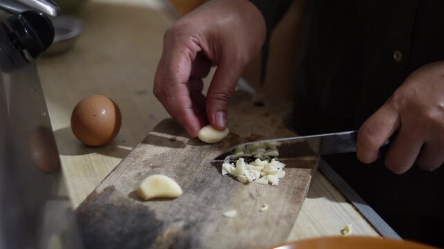 Close up of anonymous woman hands chopping and mincing fresh garlic cloves on a wooden cutting board