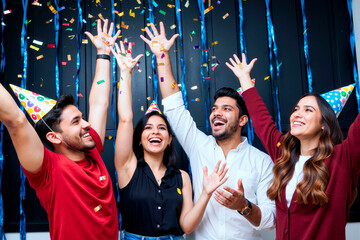 Fototapeta premium Group of young adult multiethnic friends celebrating together wearing party hats, raising hands and smiling while confetti falling, standing close and looking upward with excitement