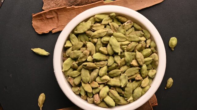 Top View of Cardamom (Elechi) in White Bowl with Cinnamon Sticks (Dalchini) on Black Background &ndash; Rotating Footage