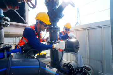 Two male industrial workers in hard hats and safety vests inspecting large pipes and valves