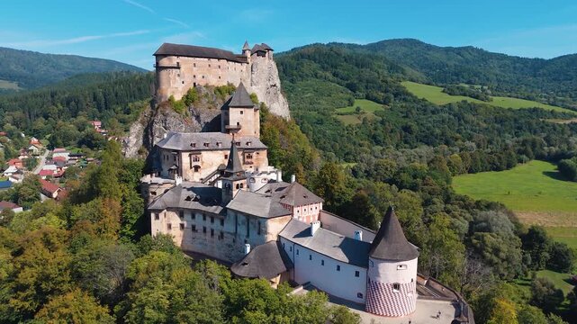Drone orbit around Orava Castle in Slovakia revealing medieval fortress perched on rocky hill above village and green mountain landscape.