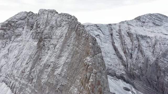 Panoramic ascending drone shot of the Throne of Zeus on Mount Olympus as Mytikas Peak emerges behind, covered in fresh snow under cloudy skies