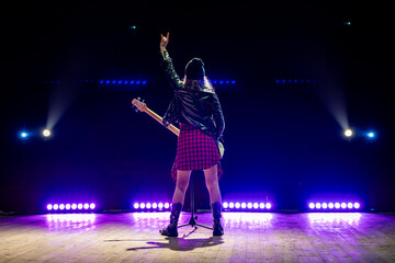 rocker girl in a black leather jacket stands with her back to the camera on stage, raising the sign of the horns hand gesture under stage spotlights. © diy13
