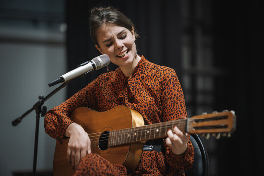 A young woman with long black hair stands against a dark background, playing an acoustic classical guitar and singing into a microphone. Classical guitar music, performing on stage.