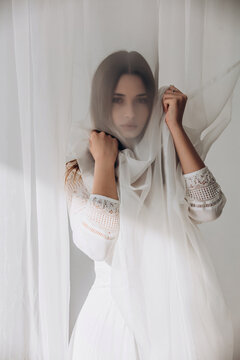 Ethereal brunette gently holding sheer white veil, soft dreamy portrait through translucent fabric.
