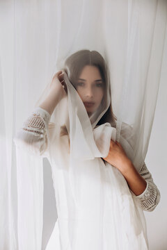 Ethereal brunette gently holding sheer white veil, soft dreamy portrait through translucent fabric.