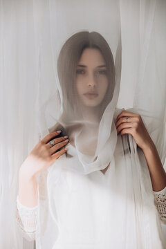Ethereal brunette gently holding sheer white veil, soft dreamy portrait through translucent fabric.