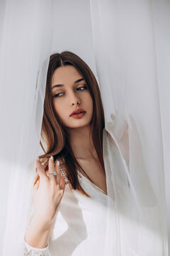 
Mysterious brunette peering through delicate white sheer curtains, soft ethereal portrait.