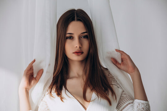 
Mysterious brunette peering through delicate white sheer curtains, soft ethereal portrait.