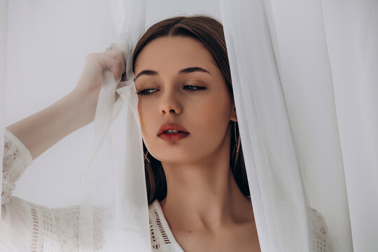 
Mysterious brunette peering through delicate white sheer curtains, soft ethereal portrait.