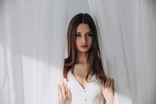 
Mysterious brunette peering through delicate white sheer curtains, soft ethereal portrait.