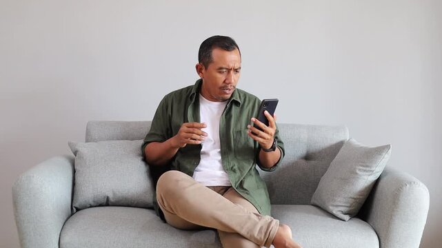 Frustrated Asian guy calling smartphone in home. Unhappy customer solve