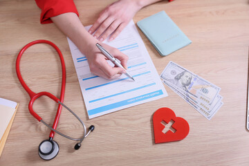 A person attentively fills out health insurance forms at a wooden desk. Nearby, a stethoscope and...