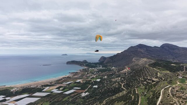 Aerial drone shot of two paragliders descending toward a valley, with mountains and the sea in the background. Scenic coastal landscape expressing adventure, freedom, and outdoor travel