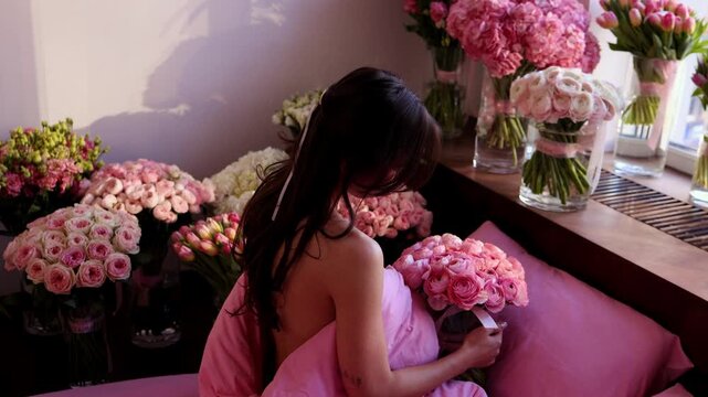 A horizontal shot from the back of a young woman sitting in a bed with pink pillows, holding a large bouquet of pink ranunculus. Surrounded by multiple flower vases.