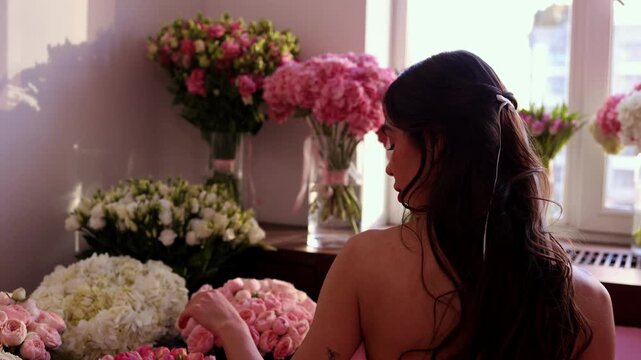 A horizontal shot from behind of a young woman with long dark hair decorated with a white ribbon. She is sitting in bed and touching numerous pink and white flower bouquets