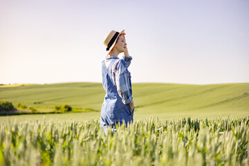 A young woman wearing overalls and a straw hat stands in a vast field of wheat, looking out over rolling green hills under a clear sky © sofiko14