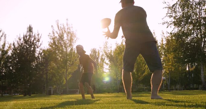 Barefoot father and son run on summer lawn. Silhouette of father playing catch with son during golden hour. Cheerful father trains boy outdoors. Lens flare sunset light and family fun. Dad and child