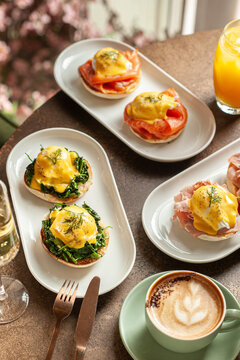 A gourmet brunch spread featuring various poached egg dishes including Eggs Royale with salmon, Eggs Florentine with spinach, and classic Benedict with ham, served with a cappuccino.