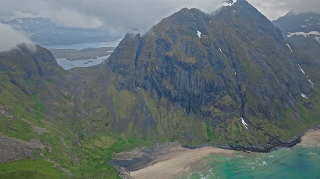 Aerial view of Kvalvika Beach tucked between tall mountains in Lofoten Islands, Norway. Norwegian drone footage showing remote sandy cove, green slopes and blue coastal waters.