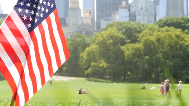 American flag on green grass in Central Park, New York, USA. Sheep Meadow, high-rise buildings and people. Skyline cityscape, Manhattan Midtown skyscrapers. United States symbol and business district.