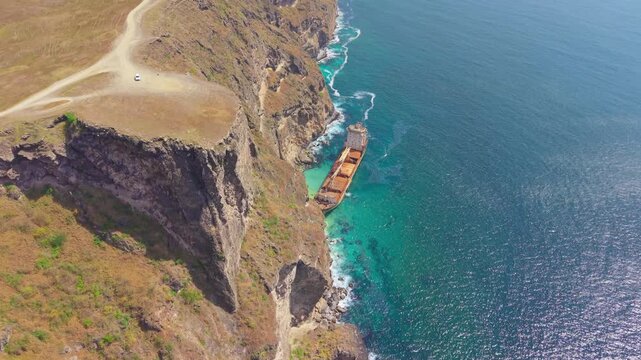 Aerial view of a grounded cargo shipwreck at the base of steep rocky cliffs along the turquoise coastline of the Arabian Sea near Salalah, Oman.