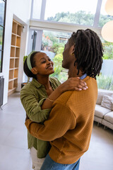 African American couple embracing at home by large windows, sofa, shelving, green headband visible