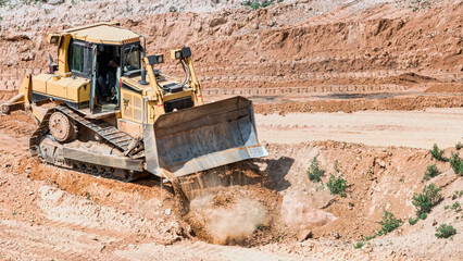A bulldozer works to clear dirt and move earth at a construction site next to a quarry. Dust rises...
