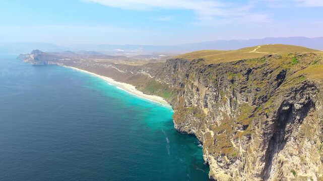 Aerial perspective of steep rocky cliffs overlooking a turquoise sea and sandy beach coastline near Salalah, Oman.