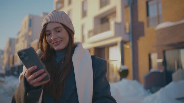 Caucasian woman walking beside stroller. Wearing knitted hat and layered coat. Holding smartphone and smiling down. Snow covering sidewalk near residential buildings. Cold season. City routine.