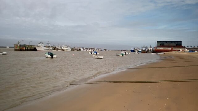 Fishing boats moored on the Guadalquivir river mouth near a sandy beach with tidal lines under a cloudy sky