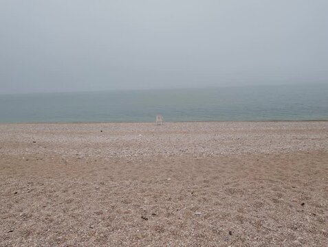 Spiaggia in una giornata nuvolosa invernale, con una sedia da bagnino bianca posizionata vicino alla riva e un mare calmo sotto un cielo grigio. Atmosfera silenziosa e contemplativa