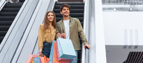A couple stands on an escalator in a mall holding shopping bags. They are both smiling and appear to be enjoying their time together. The setting is bright with modern architecture. © Prostock-studio