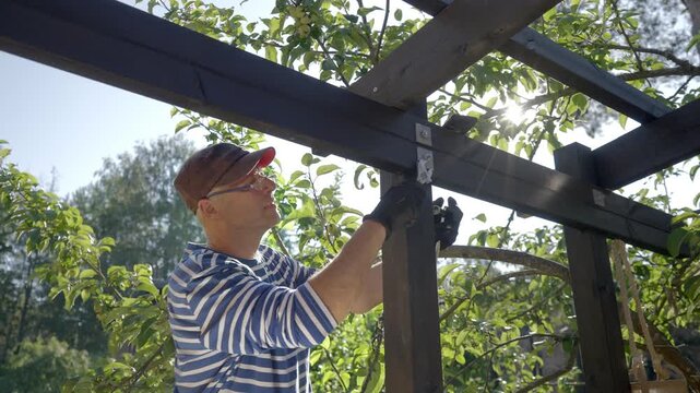 A man in a striped shirt and cap is installing a metal bracket on a wooden pergola frame in a sunlit garden. He is focused on securing the joint, working outdoors in natural light.