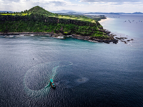 Aerial view of where the verdant headland meets the turquoise sea, a fishing boat casts its net in a circular dance of light and shadow, Tuy Hoa, Phu Yen, Vietnam.
