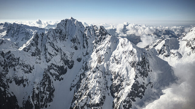 Aerial view of snow-capped mountains pierce the sky, surrounded by a sea of fluffy clouds, contrasting against the clear blue horizon, Arigna, Orobie Alps Natural park, Lombardy, Italy.