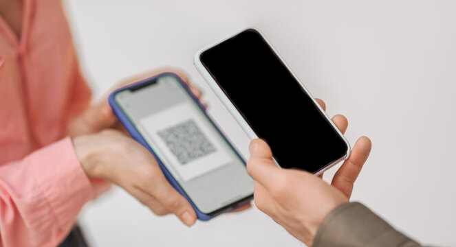 Two people hold smartphones facing each other. One screen shows a QR code. The scene likely takes place in a public or indoor setting during the day.