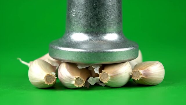 A metal garlic press slowly crushes a fresh garlic bulb, separating it into cloves against a vibrant green backdrop. Ideal for cooking and food preparation visuals.