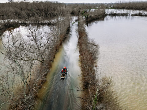Isle-Saint-Georges, France - 20 February 2026: Aerial view of a road submerged in floodwaters, with a small boat navigating the murky depths between bare trees under a grey sky.
