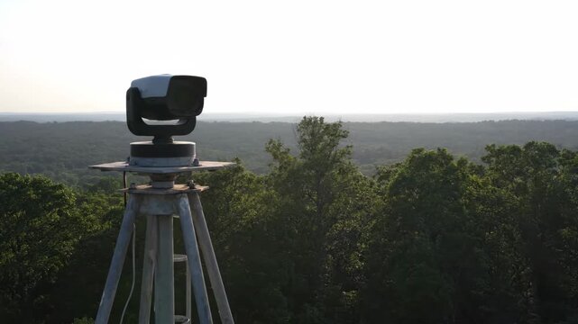 A security camera on a tripod slowly rotates, scanning the expansive green forest below under a clear sky.