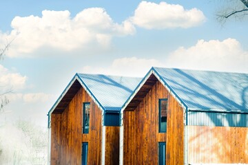 Fototapeta premium Two modern wooden houses with blue metal roofs situated under a bright sky with fluffy clouds, showcasing contemporary architectural design and natural surroundings