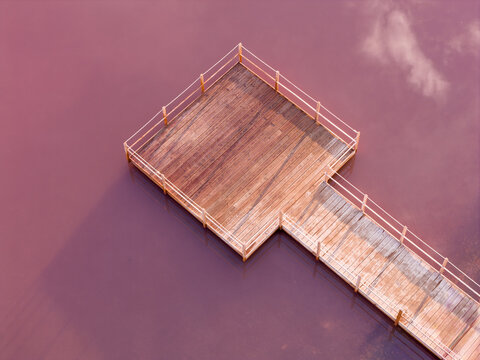 Aerial view of a wooden pier extending into the vibrant pink water of the salt marshes, the sky reflecting faintly on the water's surface, Aigues-Mortes, Occitanie, France.