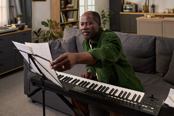 Fototapeta premium Middle aged Black man sitting on sofa playing digital piano, reaching for sheet music on music stand, practicing music in modern living room with focused expression