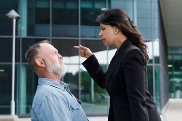 Man sits while woman speaks firmly in front of modern building in daytime