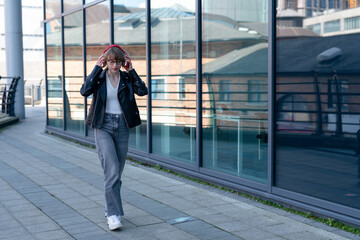 Young woman walks with headphones near glass building in city during daytime © Iryna