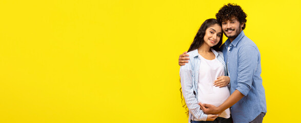 A couple stands together smiling in front of a bright yellow wall. The woman is pregnant and holds her belly. The man embraces her, showing happiness and love in their expressions. © Prostock-studio