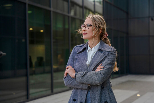 Young woman stands outside a modern building with arms crossed while looking away from the camera