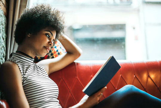 Mid adult woman with curly hair reading book while relaxing on sofa in cafe
